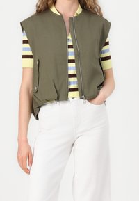 Olivgrüne ärmellose Weste mit Reißverschluss, Seitentaschen und lockerem Schnitt, getragen über einem gestreiften Kurzarmshirt. Weiße Hose.