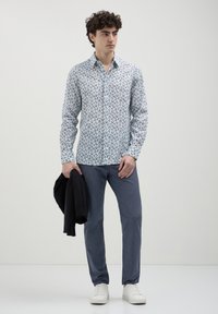 Giovane uomo che indossa una camicia bianca a fiori, pantaloni blu e sneakers bianche, con una giacca nera nella mano sinistra, in piedi su uno sfondo neutro.