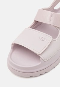 Lilla syntetisk sandaler med tykk såle, bred justerbar stropp, teksturert fotbed og UGG-logo preget på forsiden.