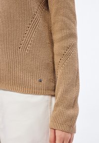 Brun strikket genser med teksturert design, med en liten logo som detalj og en avslappet passform. Ermet har en bred mansjett.