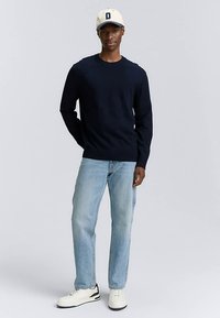 Navy plisana veste s okruglim izrezom, nošena s laganim trapericama. Bijele tenisice s crnim detaljima. Kapa ima 'D' logo u bež boji.