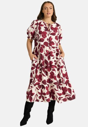 Frau trägt ein weißes Midikleid mit großem weinrotem Blumenmuster, Puffärmeln, seitlichen Taschen und schwarzen kniehohen Stiefeln.