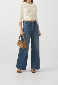 Frau trägt eine verkürzte cremefarbene Strickjacke mit goldenen Knöpfen, weite blaue Jeans, schwarze Absatzsandalen und hält eine kleine goldene Handtasche.