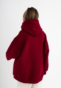 Hoodie oversize bordeaux con collo alto, cappuccio grande e spalle scese; texture morbida e polsini a coste all'orlo.