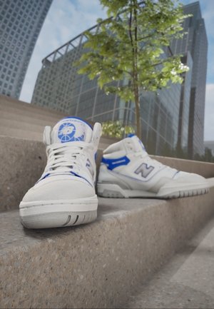Białe i niebieskie sneakersy New Balance umieszczone na betonowych stopniach z nowoczesnymi szklanymi budynkami i pełnym liści drzewem w tle.
