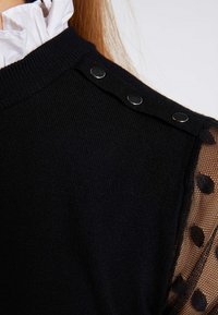 Pull en maille noire avec un col côtelé, orné de boutons décoratifs noirs sur l'épaule et des manches en mesh transparent à pois.