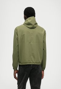 Veste à capuche vert olive avec poignets et ourlet élastiques, présentant une texture lisse et une coupe décontractée, assortie à un jean foncé.
