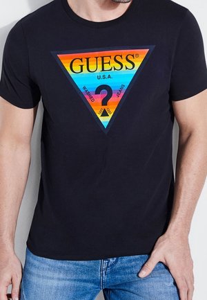 Homme portant un t-shirt noir avec un logo triangulaire multicolore indiquant « GUESS U.S.A. » et un jean bleu, debout devant un fond gris.