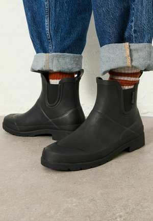 Personne portant un jean bleu retroussé, des chaussettes rayées orange et blanc et des bottes en caoutchouc noires à la cheville, debout sur un sol en béton.