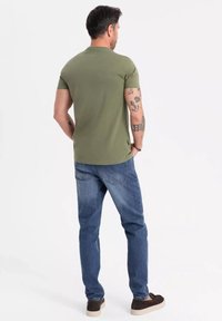 Olivově zelené tričko s krátkým rukávem a kulatým výstřihem, v kombinaci s modrými džínami a hnědými slip-on botami. Viditelné tetování na ruce.