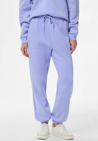 Světlé fialové tepláky vyrobené z měkké látky, s elastickým pasem se šňůrkou a lemy u kotníků. Nošeny s bílými teniskami.