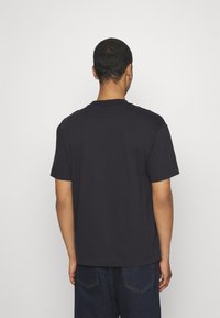 Hombre con el cabello corto, vistiendo una camiseta de color azul marino liso y jeans de mezclilla oscuros, de espaldas a la cámara, frente a una pared de color gris claro.