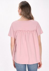 Top rosa a maniche corte con dettaglio arricciato sul retro, realizzato in tessuto morbido. Presenta un collo a barca e una vestibilità rilassata, indossato con jeans chiari.