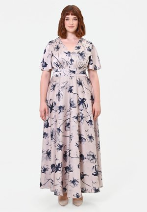 Donna con capelli rossi fino alle spalle che indossa un abito lungo floreale rosa chiaro con maniche corte e tacchi beige, in piedi davanti a uno sfondo bianco.