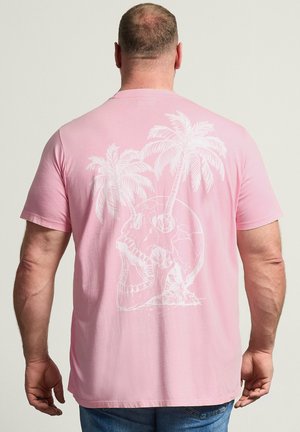 Man wearing a light pink t-shirt with a white graphic of a skull and two palm trees growing from it, viewed from the back.