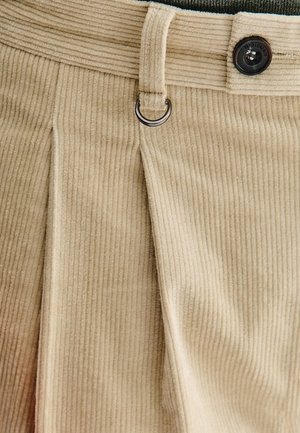 Beżowe spodnie z korduroy z pionowym prążkiem, z przyszytym fałdą z przodu, czarnym zapięciem na guzik i metalowym akcentem w formie kółka.