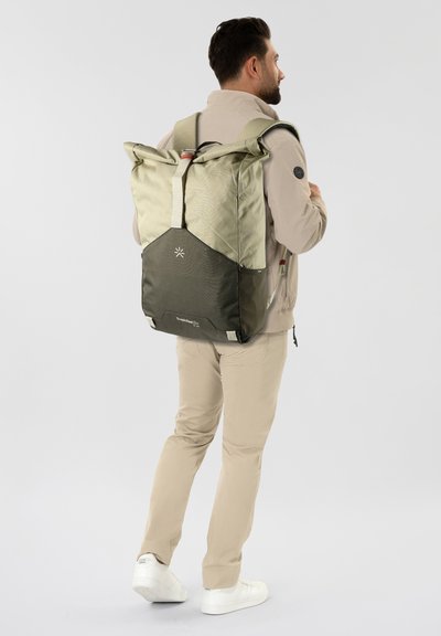 Uomo in abbigliamento beige che indossa un grande zaino a rotolo bicolore verde, in piedi contro uno sfondo grigio chiaro uniforme.