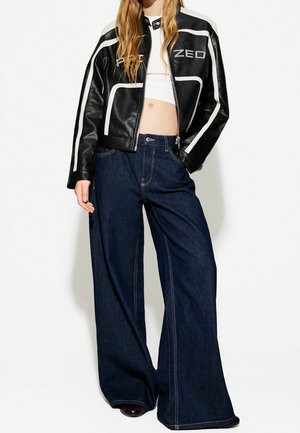 Person iført en kort sort læderjakke med hvide striber og højtaljede brede mørkeblå jeans mod en ensfarvet baggrund.