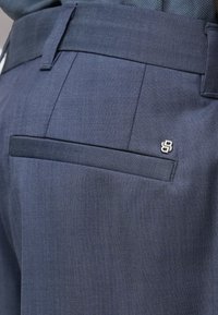 Közelkép sötétkék nadrág hátsó zsebével, övbújtatóval és a zseb széléhez közel található kis ezüst színű márkajelzéssel.