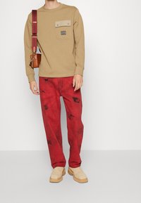 Felpa beige a maniche lunghe con tasca sul petto, abbinata a jeans rossi con tie-dye e scarpe beige slip-on. Include una piccola borsa a tracolla marrone.