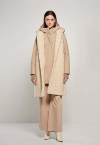 Beige buitenkleding set met een lange, fluffy crèmekleurige vest met capuchon, gelaagd over een getailleerde tan jas, gecombineerd met losse tan broeken en beige laarzen.