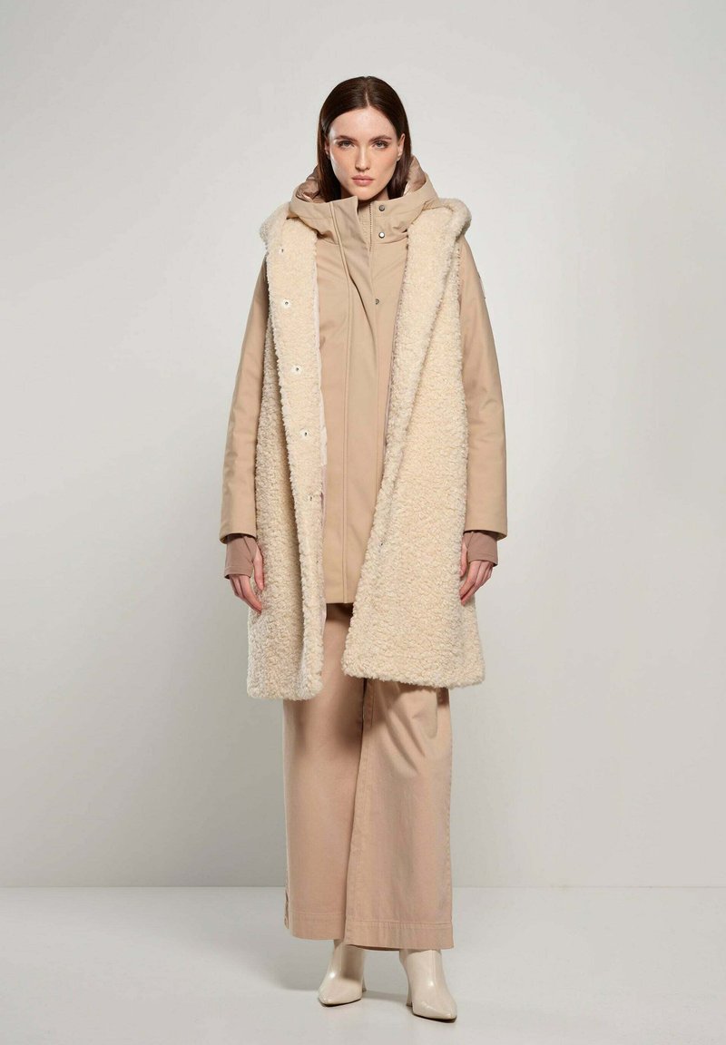 Beige buitenkleding set met een lange, fluffy crèmekleurige vest met capuchon, gelaagd over een getailleerde tan jas, gecombineerd met losse tan broeken en beige laarzen.