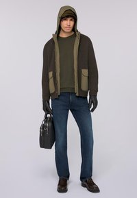 Chubasquero verde oliva con capucha, textura acanalada, dos bolsillos frontales, combinado con un suéter oscuro, vaqueros azules, guantes negros y un bolso negro.