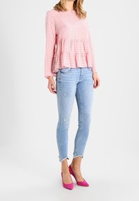 Donna che indossa una blouse a maniche lunghe in gingham rosa, jeans skinny chiari con strappi e orli sfilacciati, e tacchi alti a punta rosa brillante.