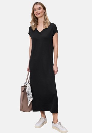 Kobieta w czarnej, midi sukience z krótkim rękawem i dekoltem w serek, w białych trampkach, trzyma torebkę w kolorze taupe z białą, wzorzystą chustą.