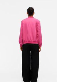 Giacca bomber rosa a maniche lunghe con polsini e orlo a costine, texture liscia. Abbinata a pantaloni neri ampi, vista da dietro.