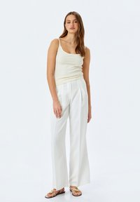 Romige tanktop en witte, geplooide wijde broek. De broek heeft subtiele verticale strepen. Het model draagt sandalen met gouden accenten.