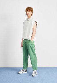 Ung person med krulligt hår som bär en ärmlös vit topp, gröna byxor, silverfärgade sneakers och ett guldhalsband, som står på ett blått golv.