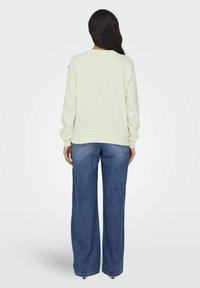 Felpa crema con maniche lunghe, polsini e orlo a costine, abbinata a jeans blu a gamba larga. Texture liscia, design semplice, senza motivi visibili.