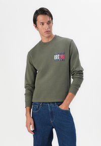 Oliven grøn sweatshirt med lange ærmer og rund halsudskæring, prydet med et logo i rød, blå og hvid på brystet. Bæres med blå jeans.