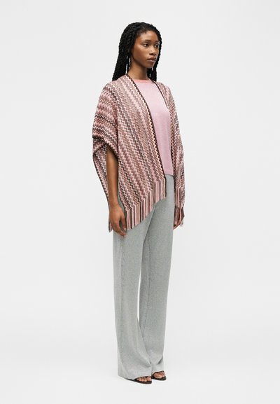 Femme debout portant un châle à motif zigzag rose sur une chemise rose, avec un pantalon gris à jambes larges et des sandales noires à bouts ouverts.