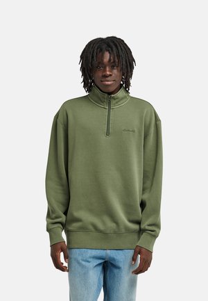 Giovane uomo dalla pelle scura con dreadlocks, indossa una felpa verde oliva con zip parziale e jeans azzurri, in piedi davanti a uno sfondo semplice.