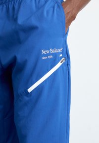 Nærbillede af en persons hånd i lommen på blå New Balance-shorts med en hvid skrå lynlåslomme og logo-teksten.