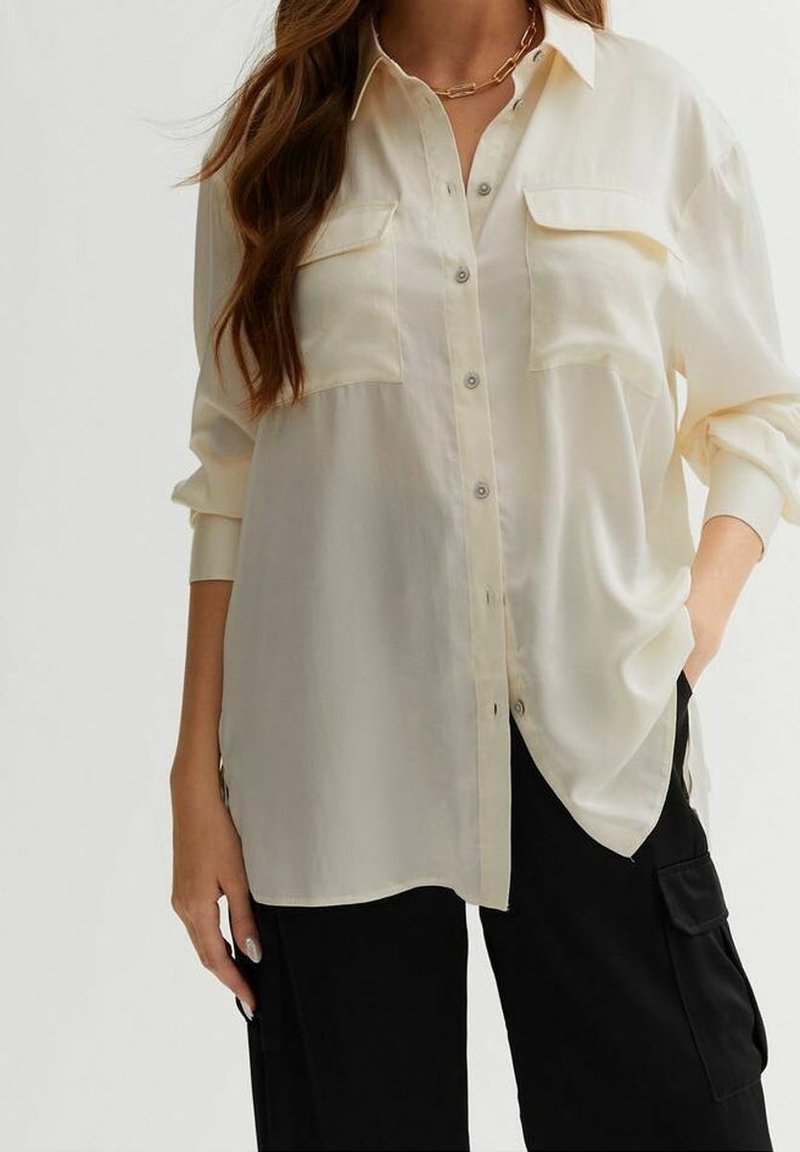 Light beige button-up shirt with two chest pockets, long sleeves rolled up, made from a smooth fabric, featuring a relaxed fit.
