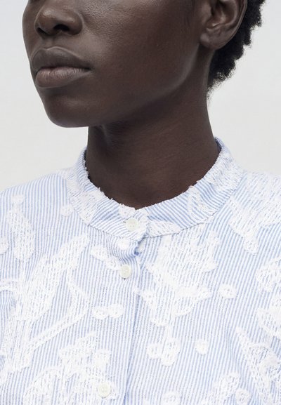 Κοντινό πλάνο ενός ατόμου που φοράει μπλε και άσπρο ριγέ πουκάμισο με υφασμένους λουλουδένιους σχεδιασμούς και μανδαρινένιο γιακά με λευκά κουμπιά.