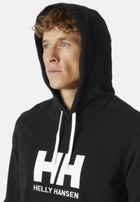 Černá mikina s kapucí a šňůrkami, s velkým bílým logem "HH" a textem "HEL"LY HANSEN". Hladká textura, neformální design, detailní pohled.