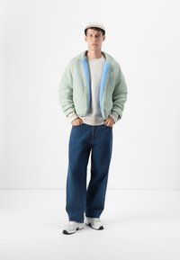 Jonge man die staat en een lichtgroene jas met blauwe voering, een beige pet, een sweatshirt, brede blauwe jeans en witte sneakers draagt tegen een witte achtergrond.