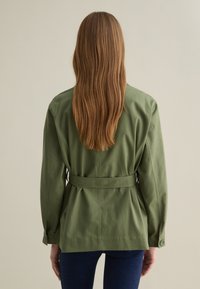 Giacca verde oliva con cintura, maniche lunghe, vestibilità rilassata e polsini con bottoni. Tessuto liscio senza motivi visibili o accessori.