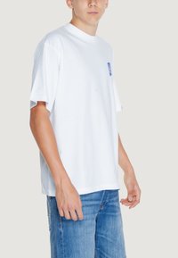 Camiseta de algodón blanca con mangas cortas y cuello redondo. Presenta un diseño gráfico azul en el lado izquierdo del pecho. Corte recto clásico.