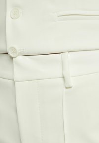 White fabric close-up featuring two buttons, a waistband with a loop, and a side pocket. The texture appears smooth with fine stitching details.