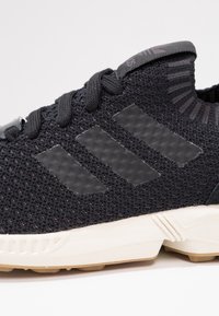 Zwarte gebreide sportshoe met drie gestructureerde zwarte strepen, rubberen accenten en een witte midsole. Heeft bijpassende zwarte veters en kraag.