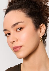 Złota okrągła kolczyk, z teksturowaną powierzchnią. Osoba ma kręcone ciemne włosy, neutralne usta, gładką skórę i zielono-brązową bluzkę.