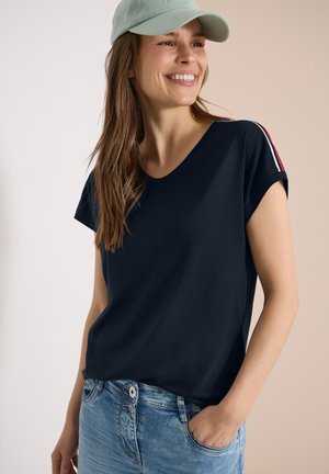 Lächelnde Frau mit hellgrüner Kappe, dunkelblauem T-Shirt mit roten und weißen Streifen auf den Schultern und hellblauen Jeans, eine Hand in der Tasche.