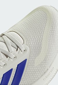 Zapatilla deportiva gris claro con textura de malla, que presenta franjas de acento azules, cordones planos y la marca CLOUDFOAM en la suela.