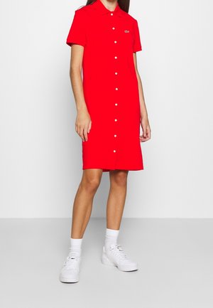 Žena v červených košilových šatech délky ke kolenům s krátkými rukávy, doplněná bílými ponožkami a robustními bílými teniskami.