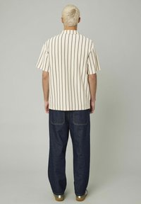 Chemise à manches courtes crème avec des rayures verticales noires, associée à un pantalon en denim sombre ample et des chaussures rehaussées de vert.